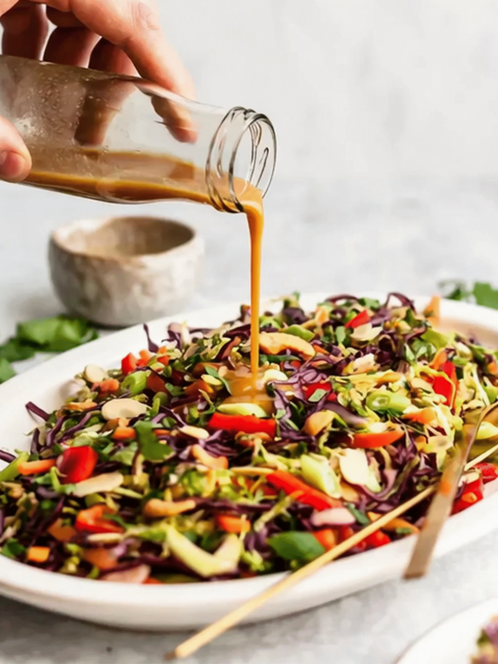 Pouring sesame ginger dressing over a colorful Asian-style salad with red cabbage, bell peppers, and peanuts