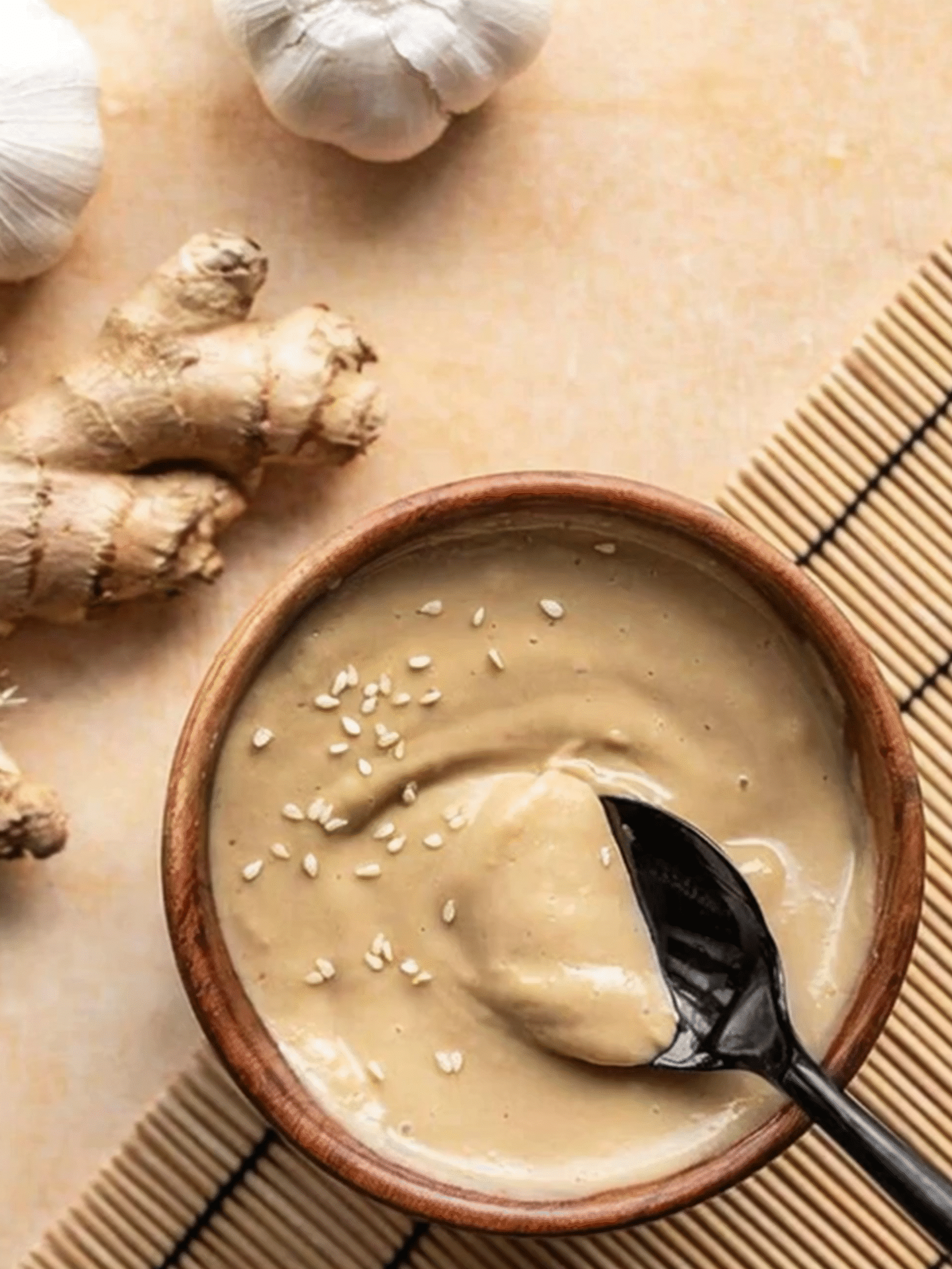 Thick sesame ginger dressing in a wooden bowl with a spoon, garnished with sesame seeds, surrounded by fresh garlic and ginger