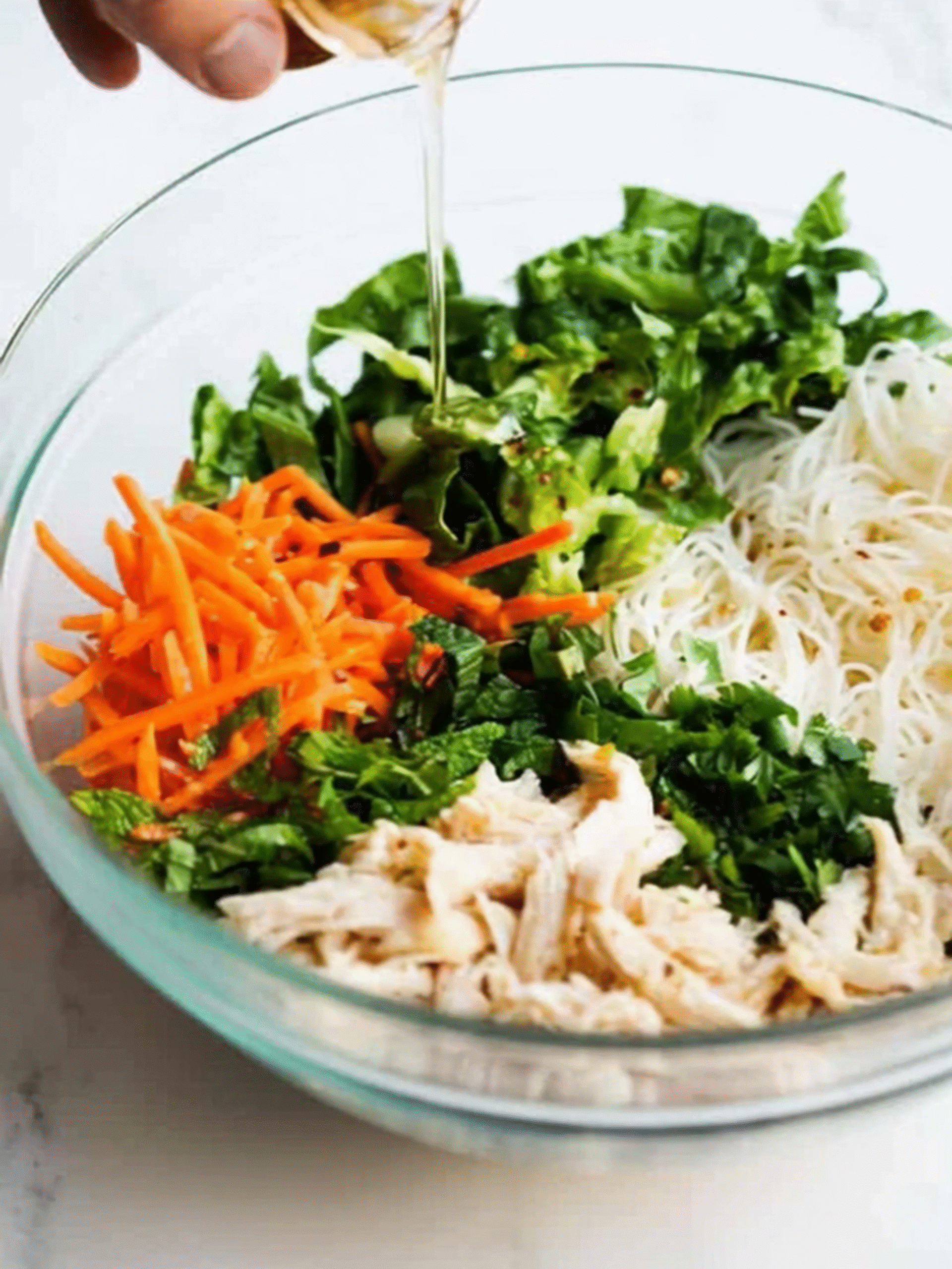 Adding Dressing to Vietnamese Noodle Salad with Fresh Herbs Pouring dressing over Vietnamese Noodle Salad with shredded chicken, noodles, carrots, herbs, and lettuce in a glass bowl