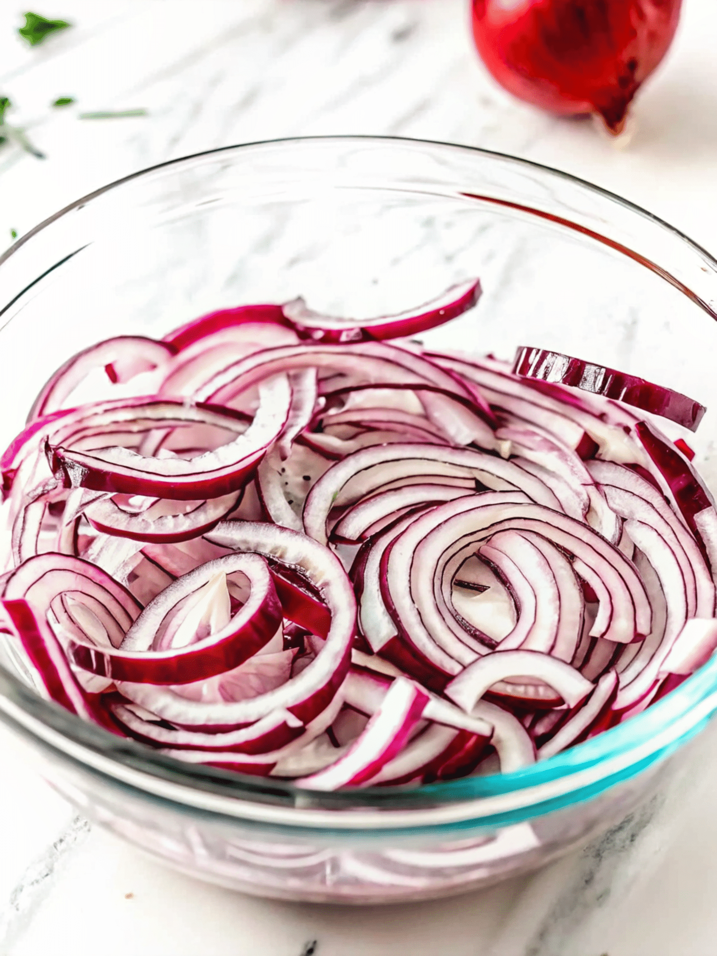 Soaking Red Onions for Tomato Cucumber Avocado Salad Thinly sliced red onions soaking in a clear glass bowl on a marble countertop