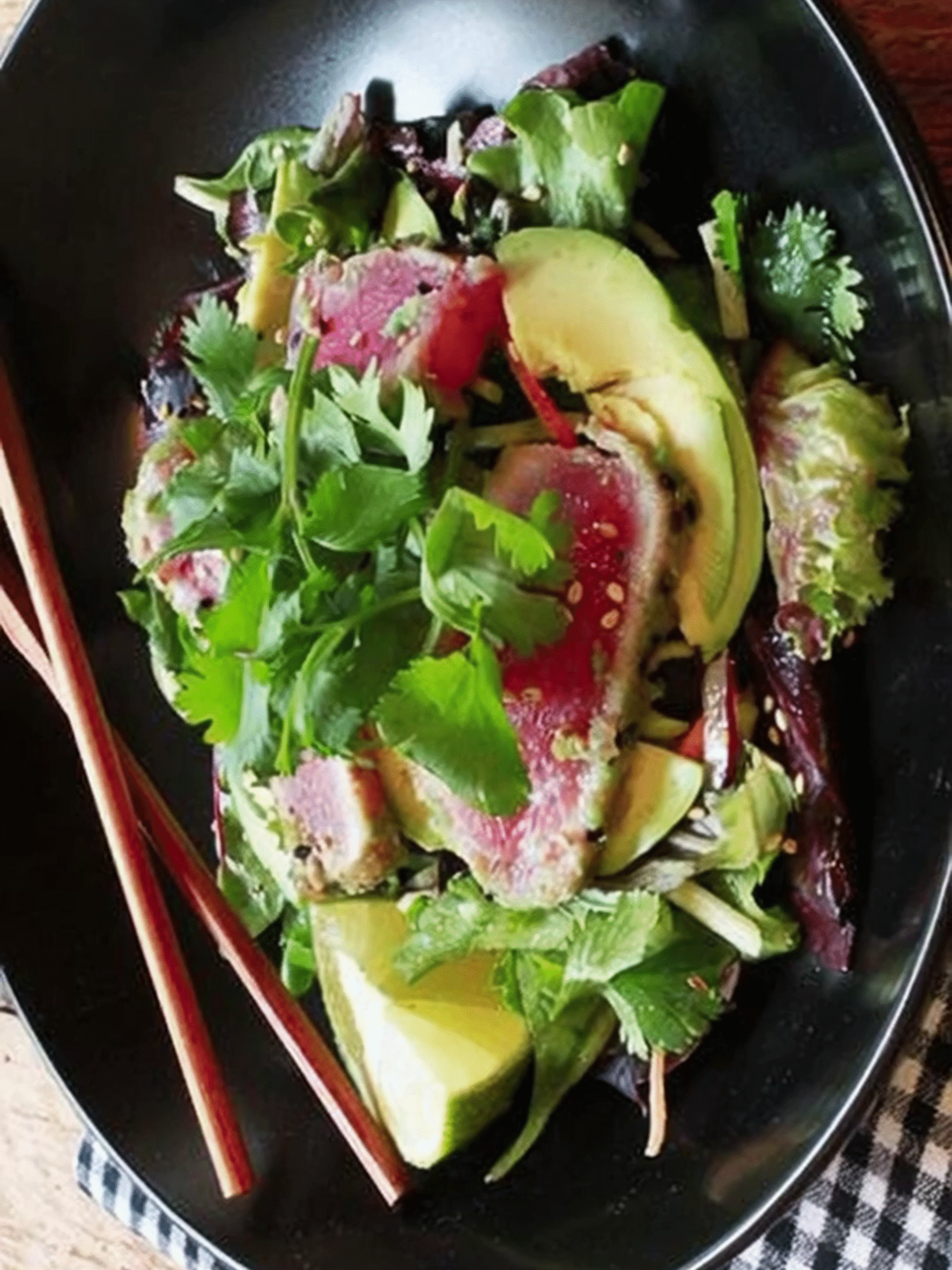 Seared tuna salad with avocado, fresh greens, lime wedges, and miso dressing in a black bowl