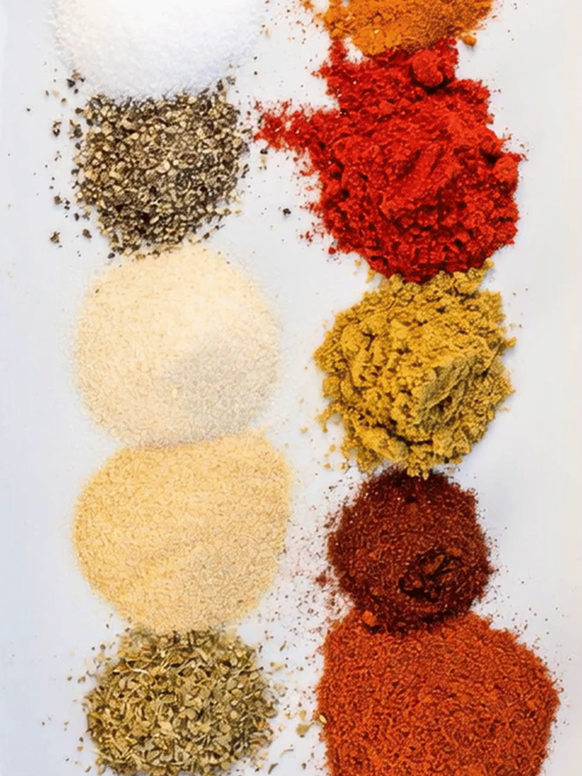Colorful Spice Mix for Pink Salt Seasoned Roasted Chicken Assorted colorful spices including salt, black pepper, paprika, turmeric, cumin, garlic powder, onion powder, and oregano arranged neatly on a white surface.