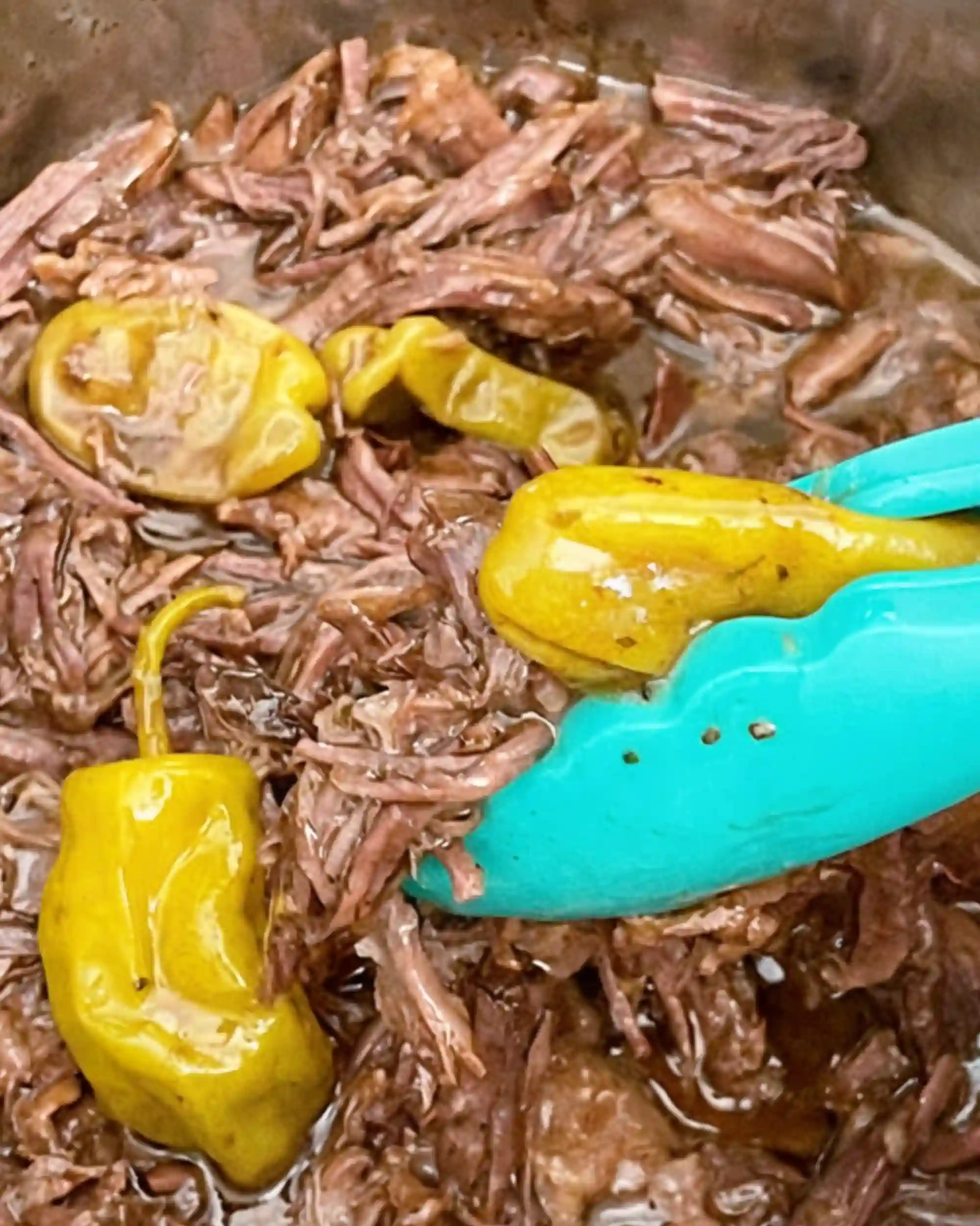 Shredded Mississippi Pot Roast in gravy with pepperoncini peppers being lifted by turquoise tongs