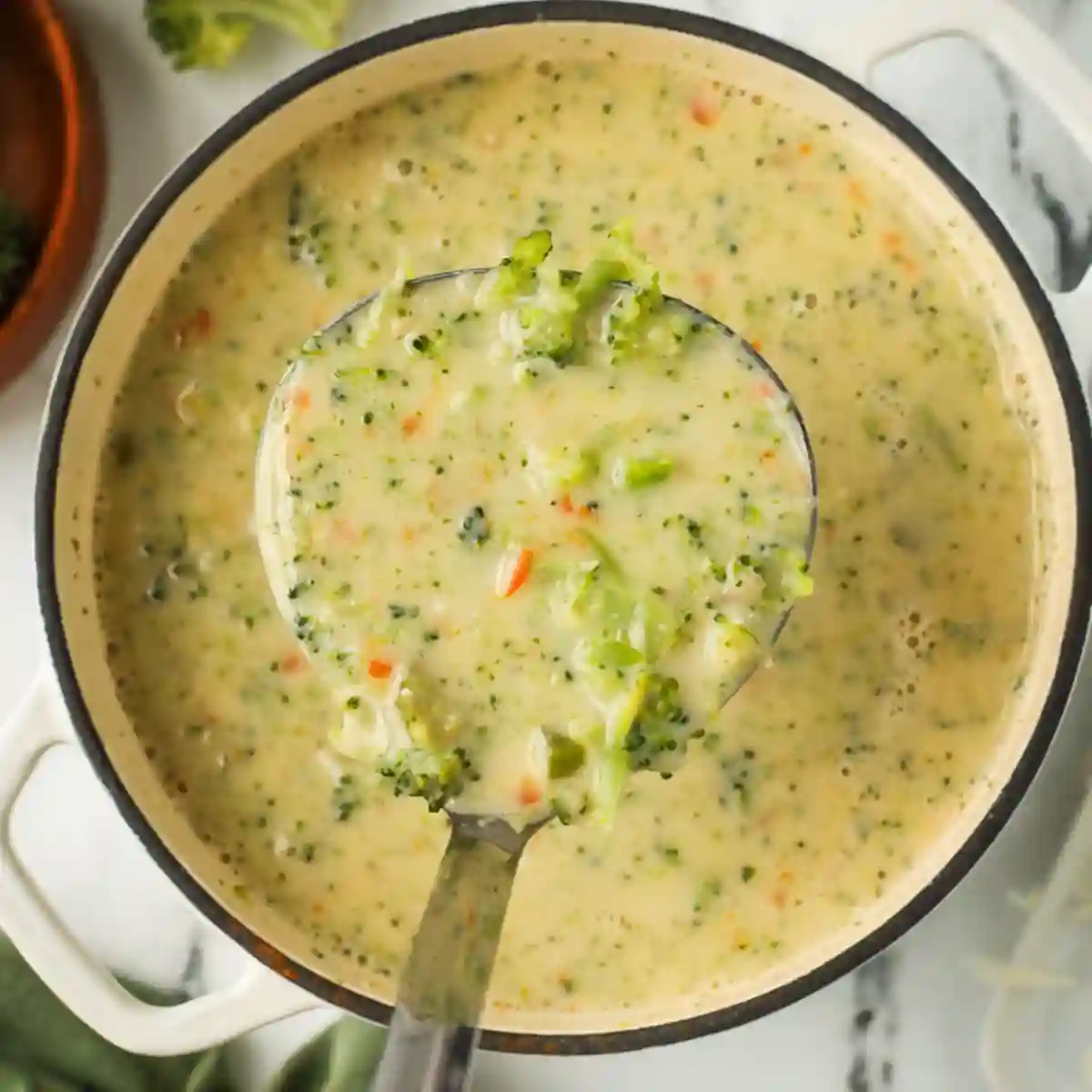 High protein broccoli cheddar soup in a white pot with a ladle full of creamy soup showing chunks of broccoli, carrots, and cheese.