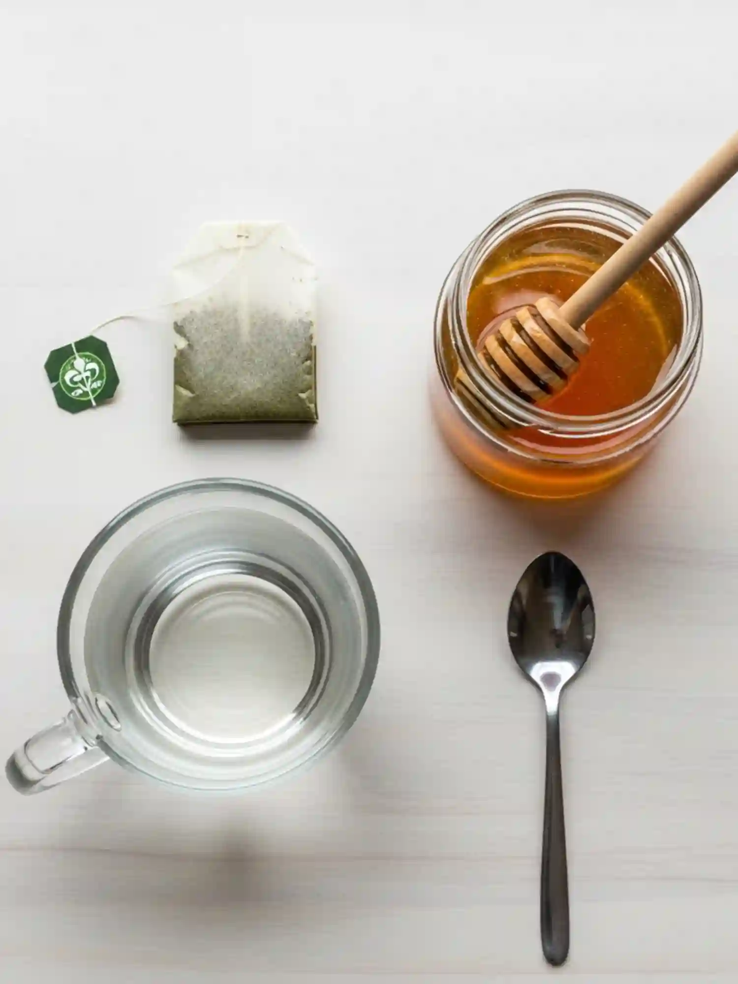 Flat lay of green tea ingredients including a tea bag, honey jar with dipper, glass mug of water, and a spoon on a light background.