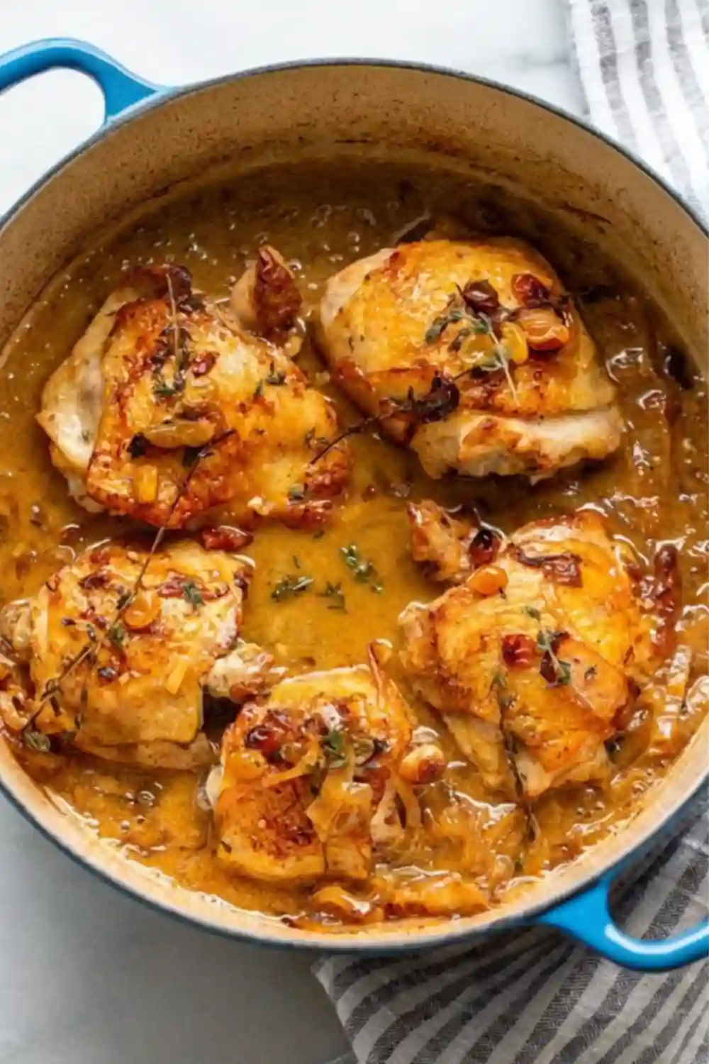 Golden apple cider braised chicken thighs simmered in a rich sauce with onions and golden raisins in a blue Dutch oven