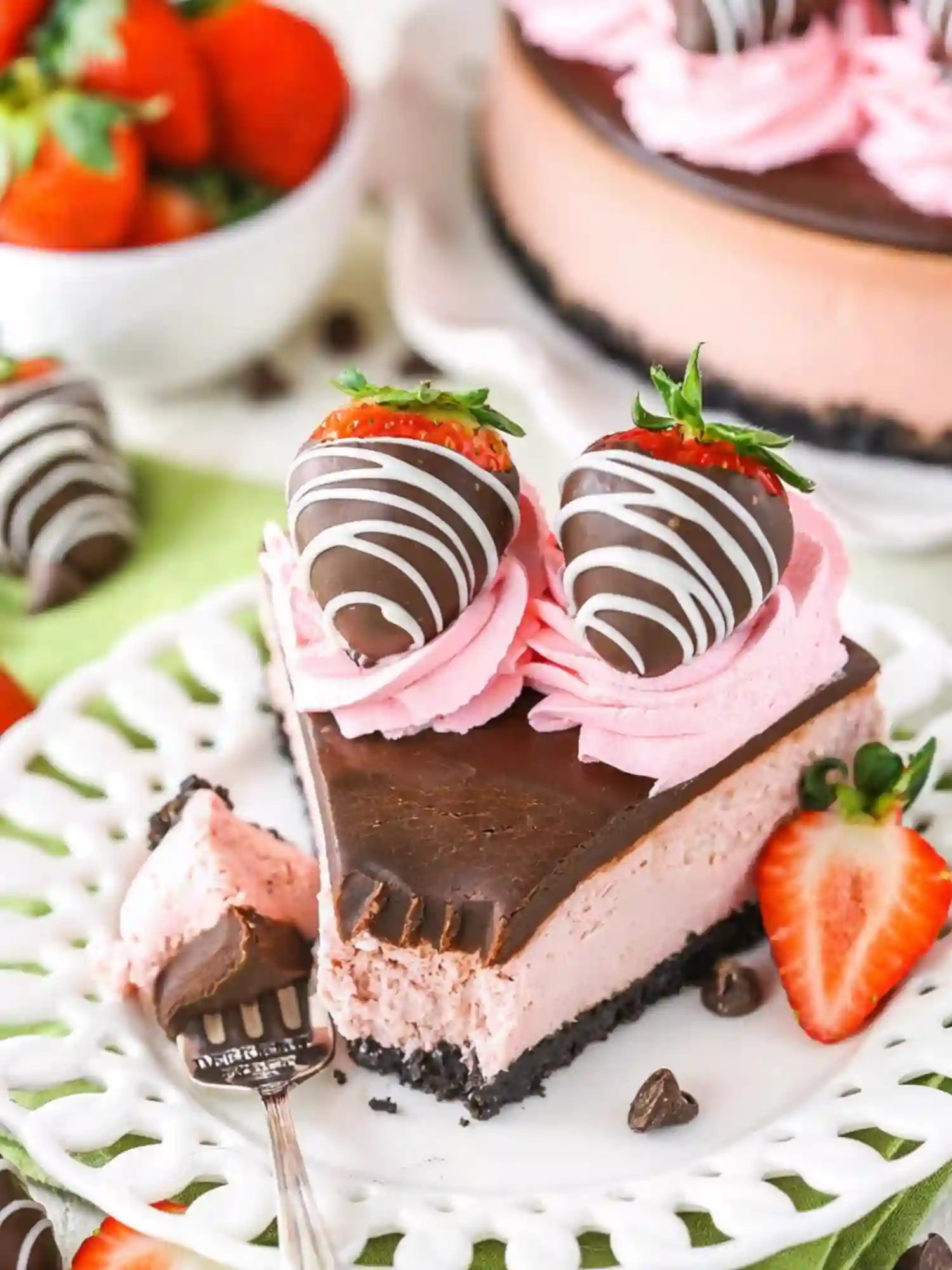 Slice of chocolate covered strawberry cheesecake with pink whipped cream, chocolate ganache, and two chocolate-dipped strawberries on top.