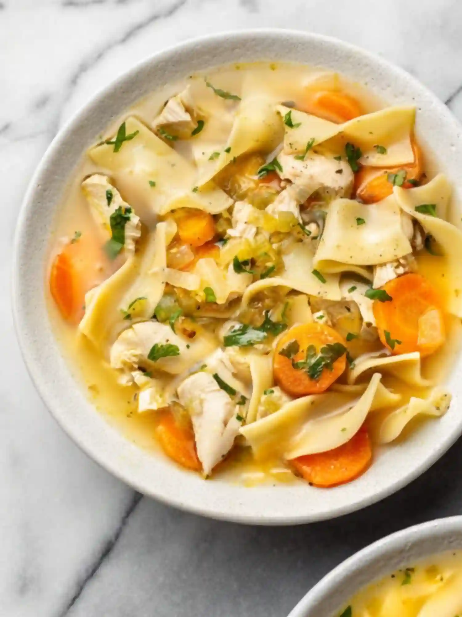 Bowl of chicken noodle soup with egg noodles, carrots, shredded chicken, and fresh parsley on a marble surface