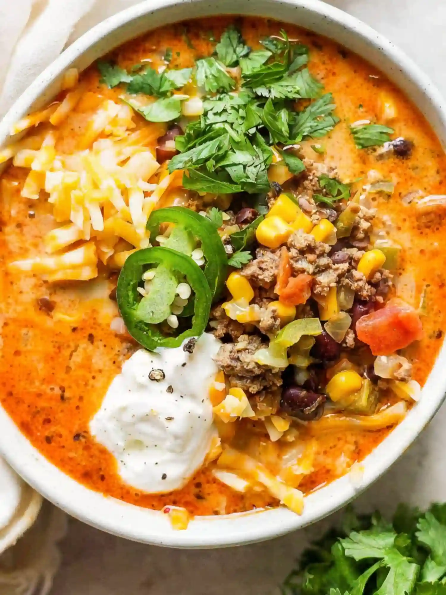 Bowl of creamy taco soup topped with sour cream, shredded cheese, sliced jalapeños, ground beef, corn, black beans, and fresh cilantro.