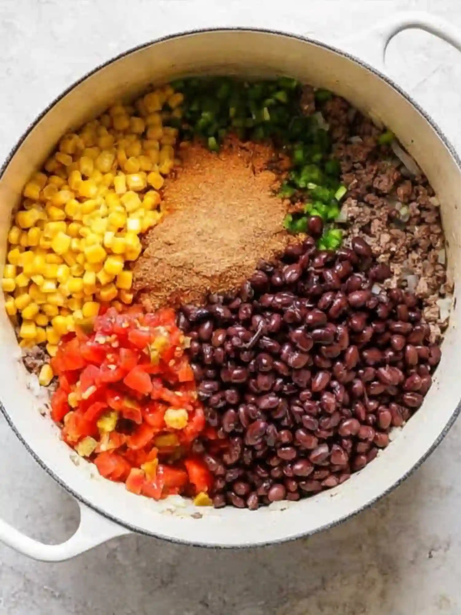 Large white pot filled with layers of corn, black beans, diced tomatoes, taco seasoning, peppers, and ground beef before stirring.