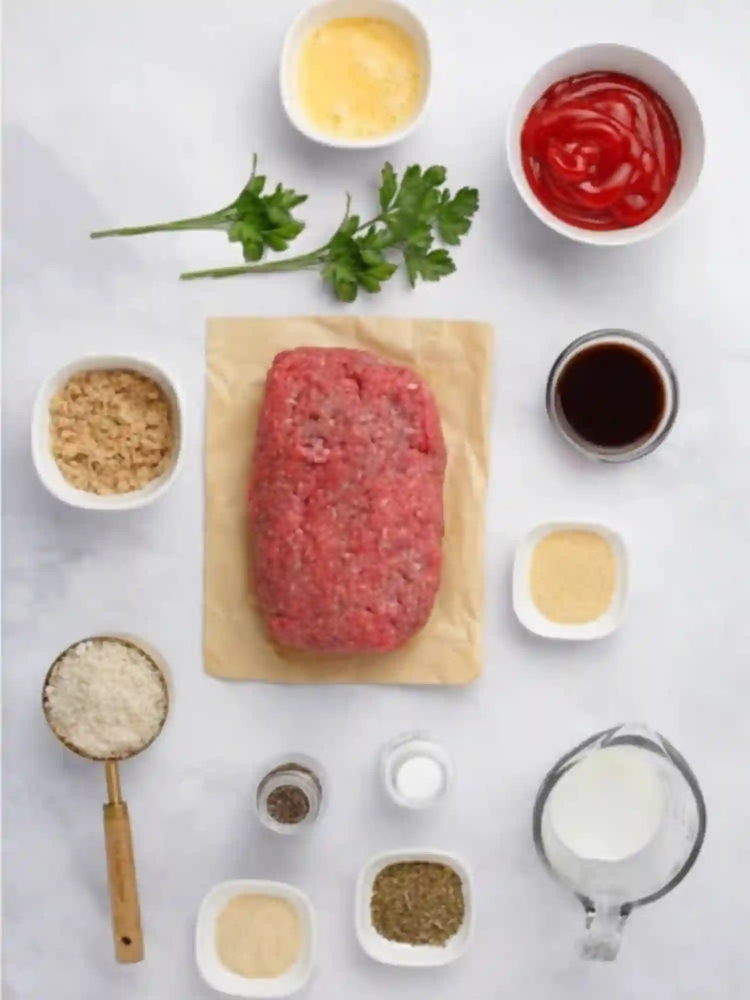 Flat lay of meatloaf ingredients including ground beef, eggs, ketchup, parsley, breadcrumbs, Worcestershire sauce, seasonings, and milk on a white background