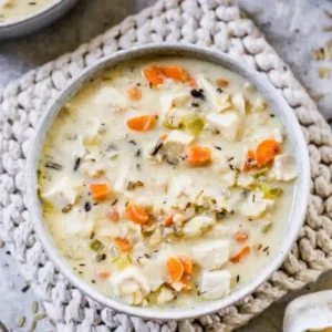 Bowl of creamy chicken and wild rice soup with carrots, celery, and herbs served on a textured placemat.