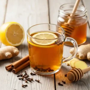 Glass mug of clove tea recipe with a lemon slice, surrounded by fresh ginger, cinnamon sticks, honey jar, and whole cloves on a rustic wooden surface.