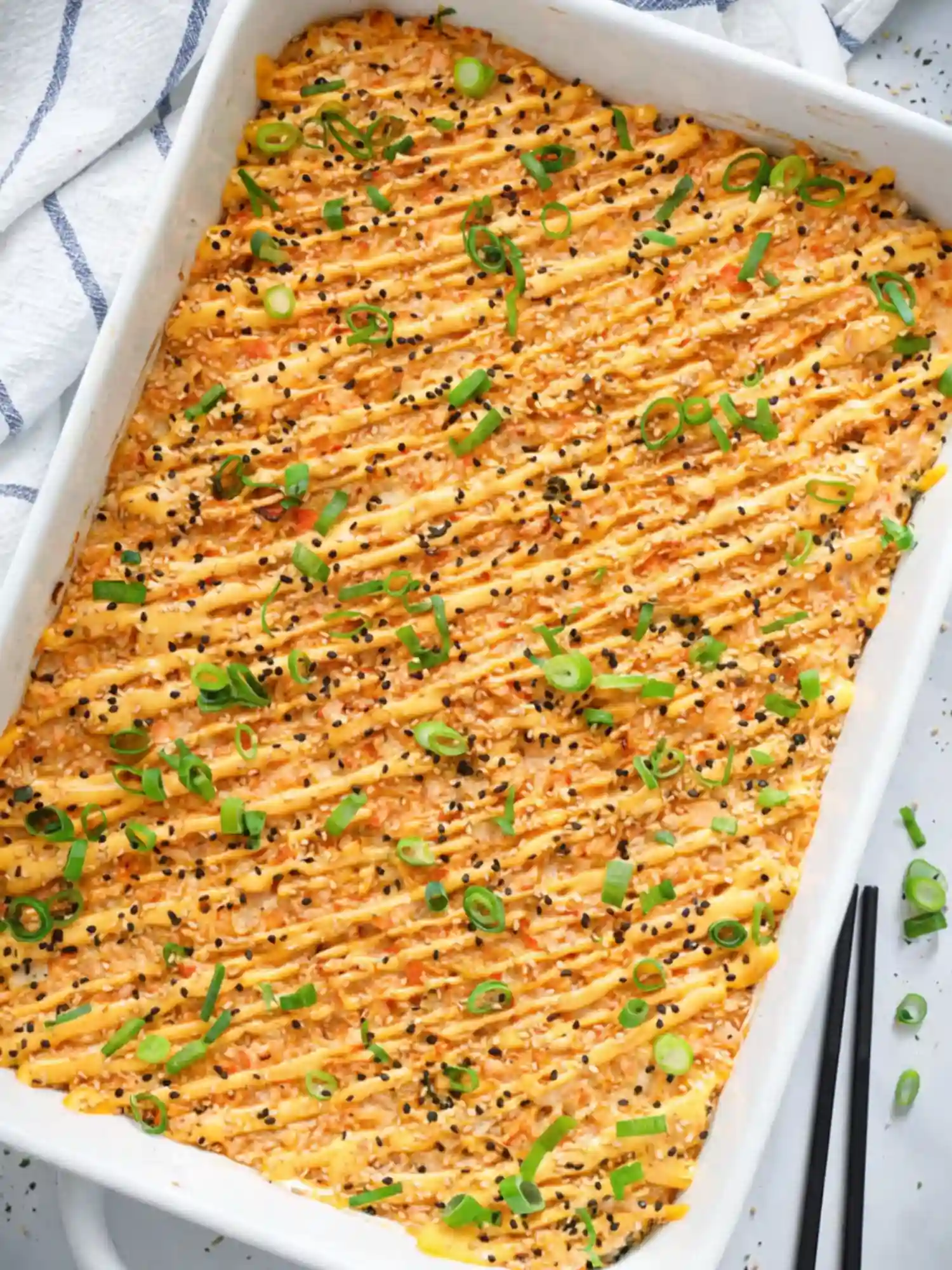 Baked Salmon Sushi Casserole with Spicy Mayo and Green Onions Overhead shot of a freshly broiled salmon sushi bake in a white rectangular baking dish, drizzled with spicy mayo and topped with sliced green onions and sesame seeds.
