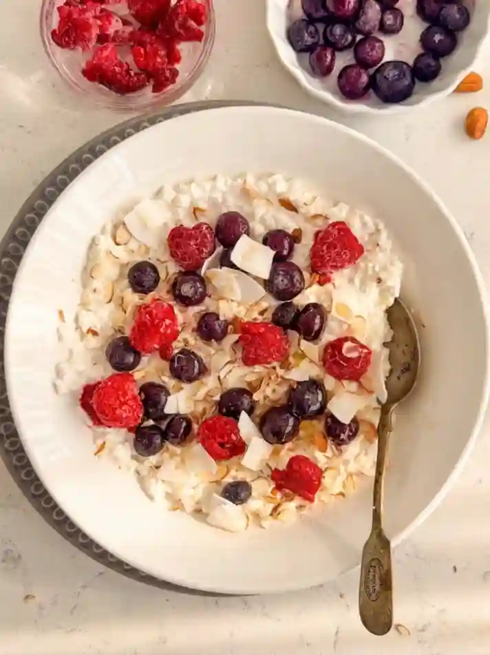 Cottage cheese breakfast bowl topped with frozen raspberries, blueberries, coconut flakes, and almonds in a white bowl with spoon