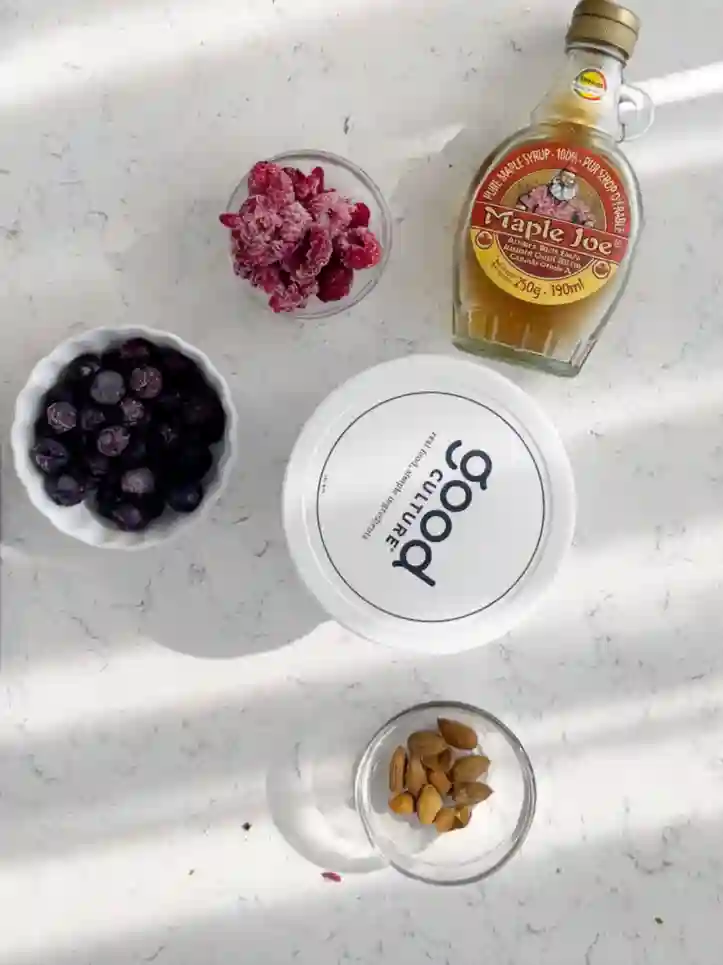 Ingredients for a high protein breakfast cottage cheese bowl including cottage cheese, maple syrup, almonds, raspberries, and blueberries