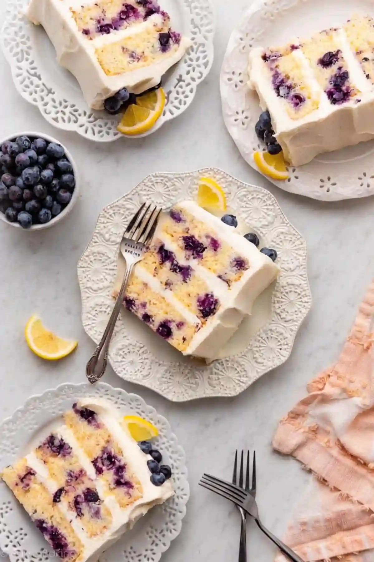 Plated slices of lemon blueberry cake with cream cheese frosting, garnished with lemon wedges and blueberries, served on elegant white dishes.