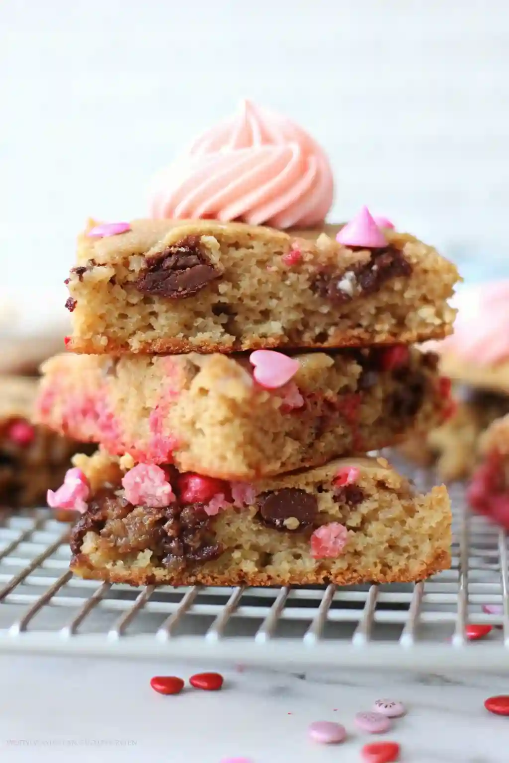 Heart Shaped Cookie Cake Slices with Frosting Stacked slices of heart shaped cookie cake with visible chocolate chips, pink sprinkles, and piped frosting on top.