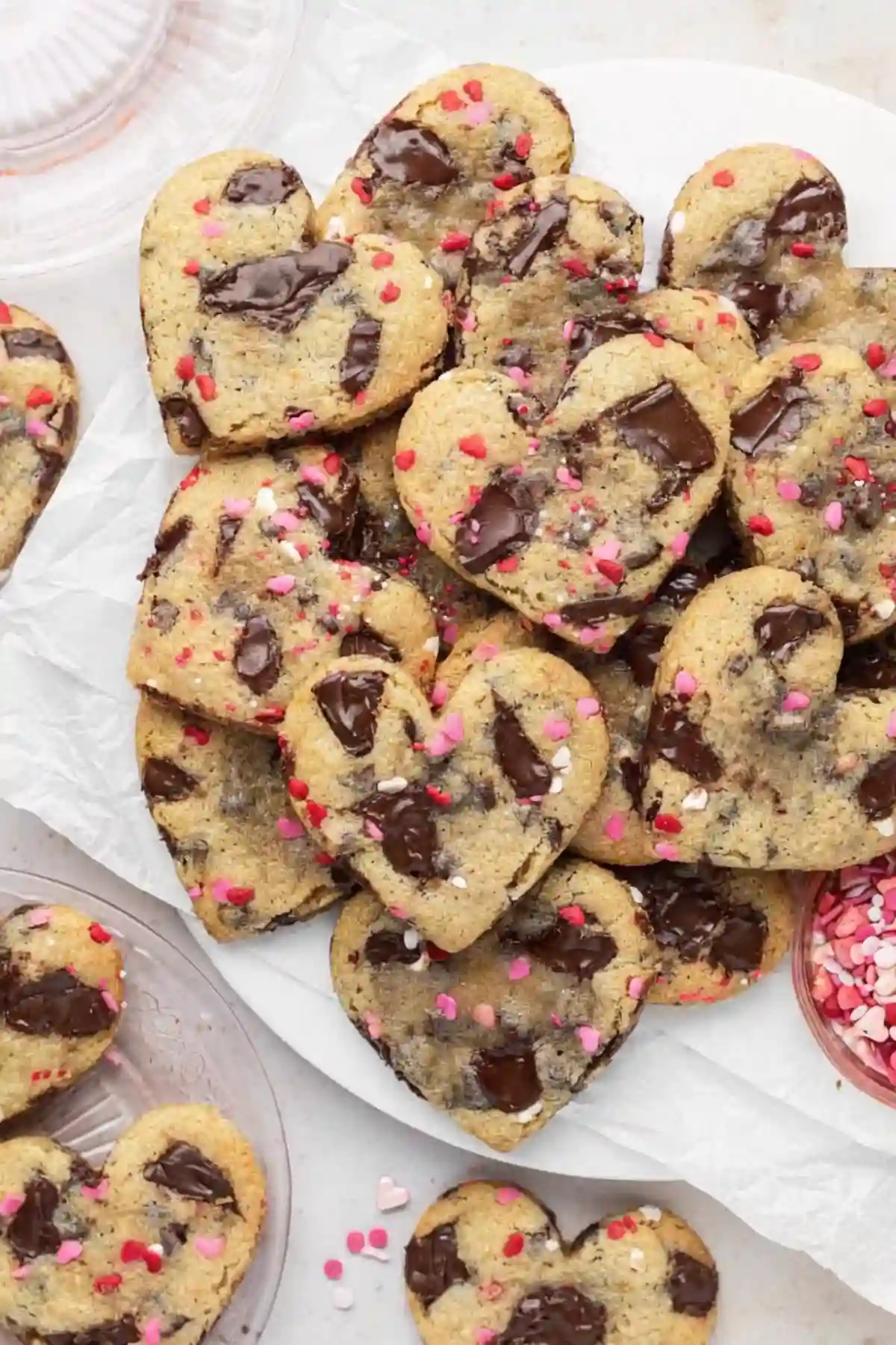A plate full of heart shaped chocolate chip cookies with melted chocolate chunks and colorful Valentine’s sprinkles, arranged on parchment paper