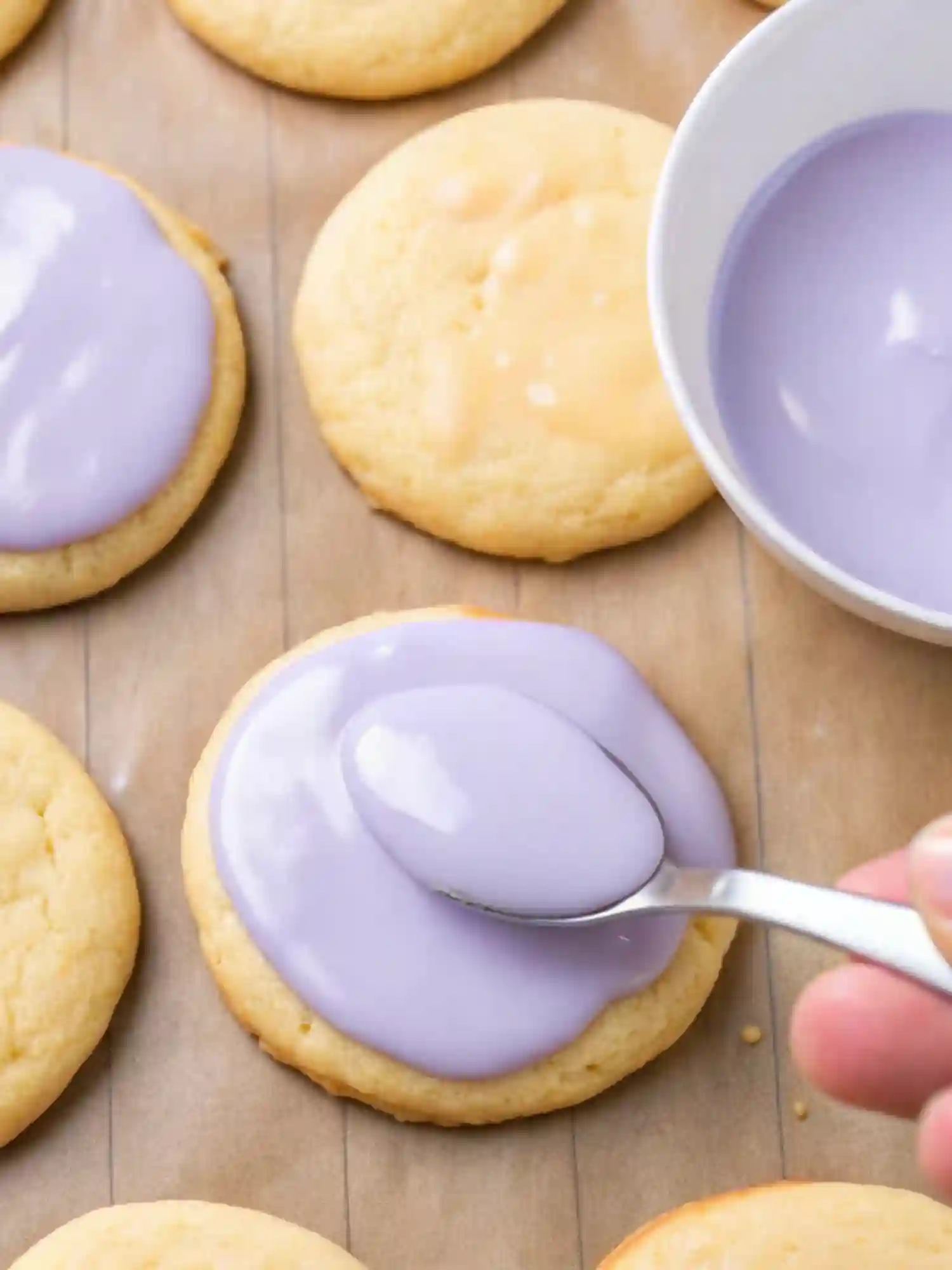 A hand spreading purple glaze onto lemon lavender cookies with a spoon over parchment paper.