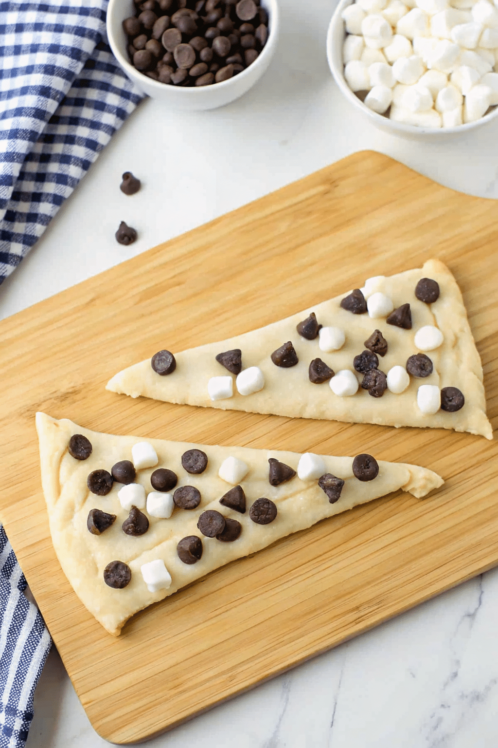 Two unbaked crescent dough triangles topped with mini marshmallows and chocolate chips on a wooden board, ready to roll
